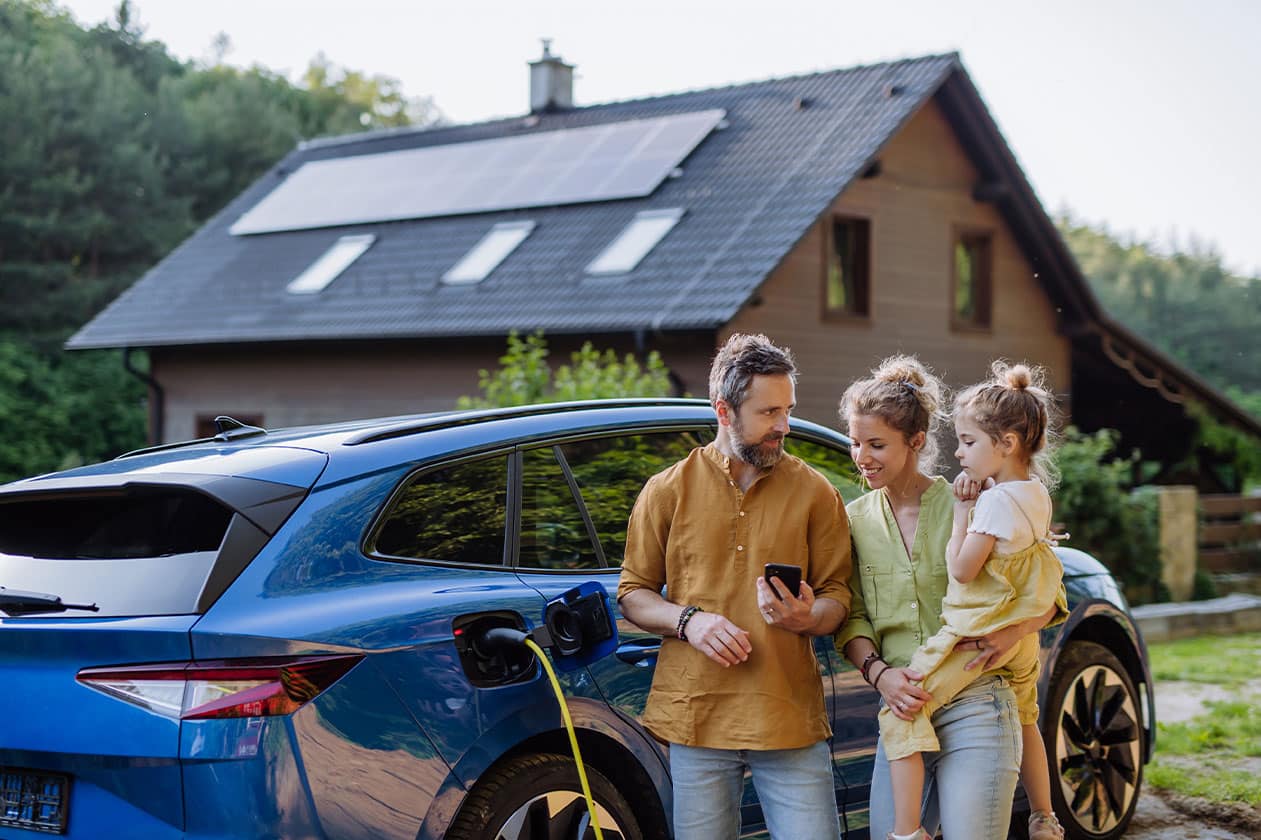Junge Familie vor E-Auto und Haus mit Photovoltaik steht für Nachhaltigkeit